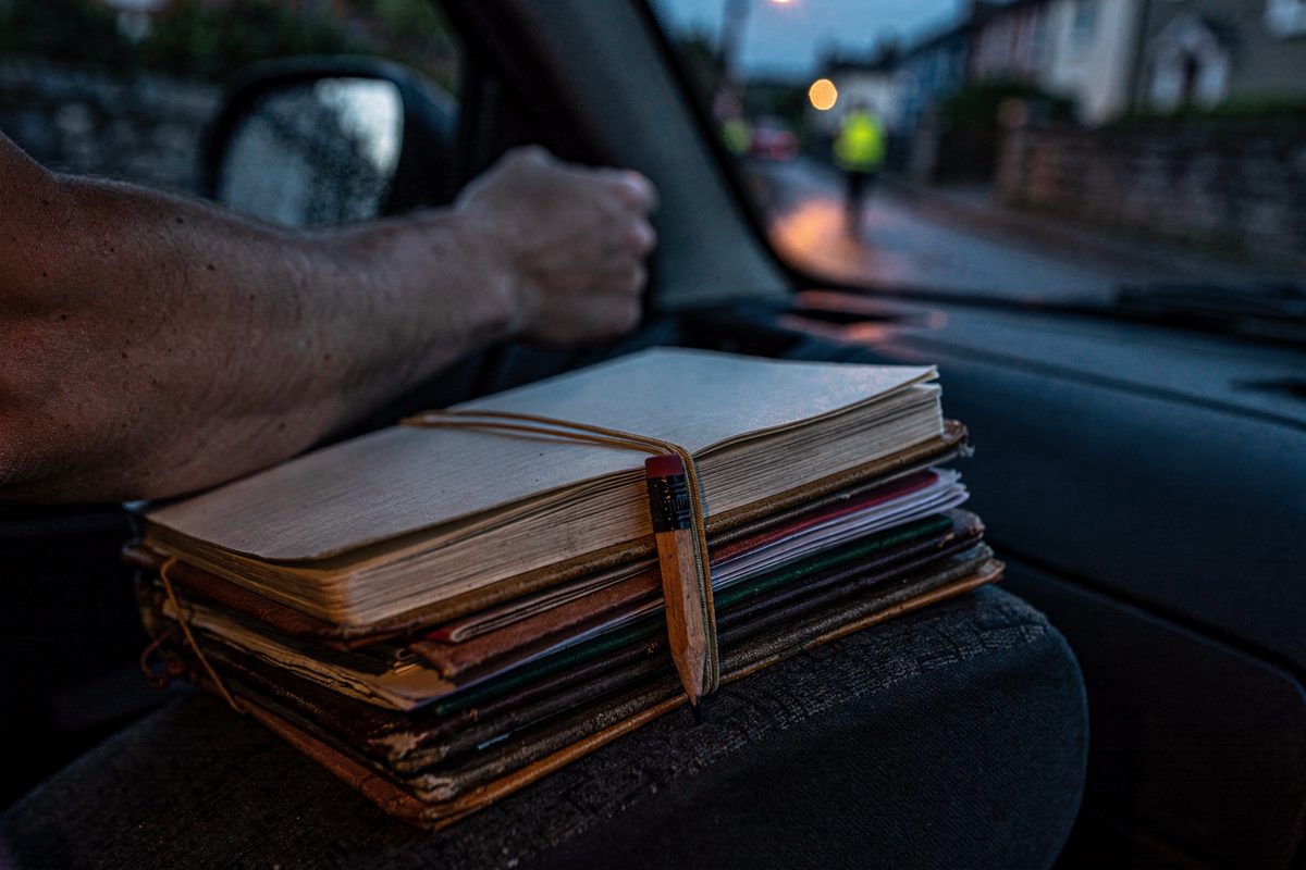 Close detail at dusk inside a parked hatchback, a thick bundle of filled pocket notebooks held together with elastic bands on the passenger seat, a blunt.