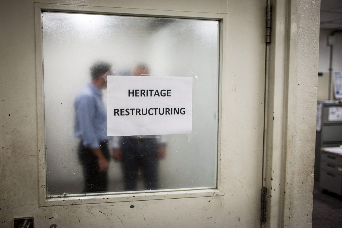 Mid-morning fluorescent light in a factory office corridor, a temporary printed sign reading "HERITAGE RESTRUCTURING" taped to a glazed door, two figures.