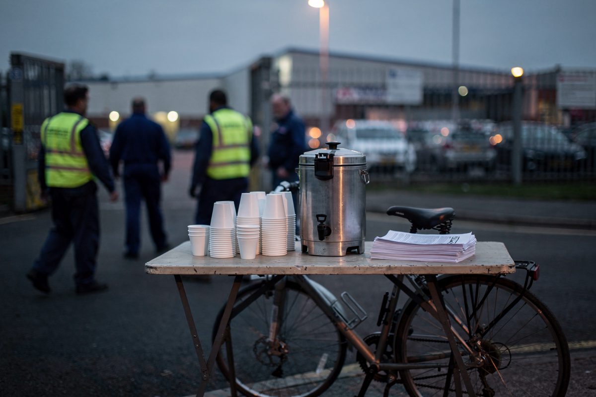 Dawn at a car plant gate in flat grey-blue light, a union trestle table with a tea urn and stacked polystyrene cups, workers in overalls crossing the road.