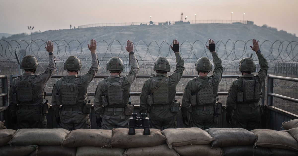 Dawn light over a hillside observation post beside a razor-wire border fence, a row of soldiers shot from behind raising their right arms in unison toward the.