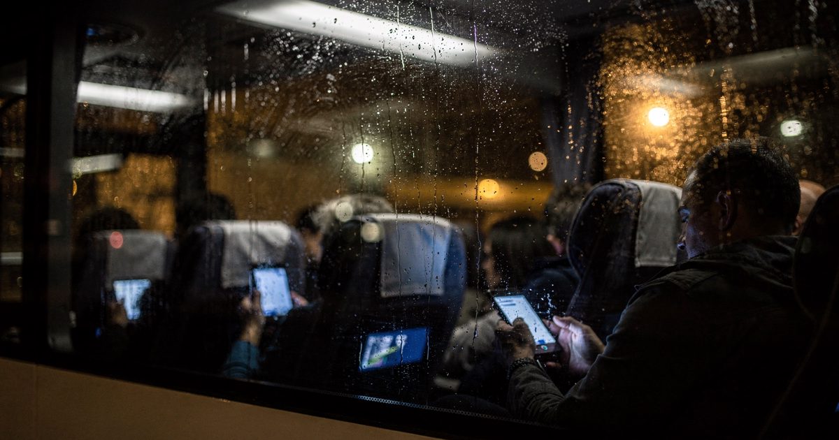 A tight, dimly lit shot through the rain-streaked window of a parked motorcoach, showing the blurred silhouettes of exhausted passengers hunched over their.