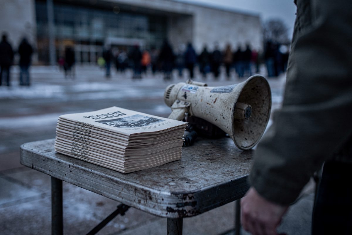 Close detail in cold early-morning light on a Washington arts plaza, a stack of folded paper season programmes on a trestle table weighted down by a battered.