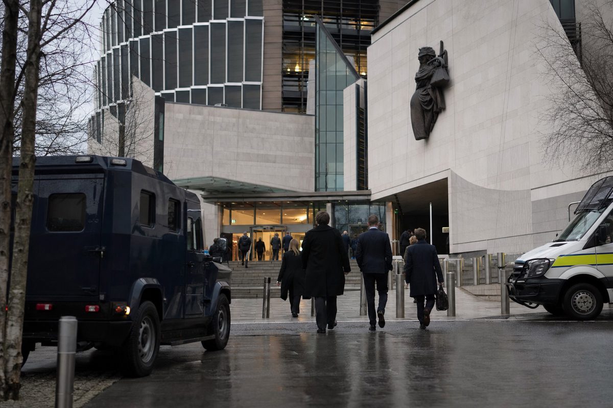 A press photography scene outside a modern government justice building in Dublin under grey skies, security vehicles near the entrance and a small group of.