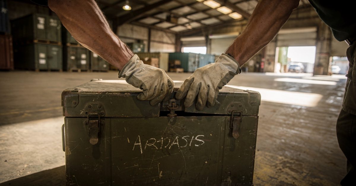Close-up of gloved hands prying open the latch of a dented olive metal footlocker inside a hot storage shed, harsh midday light through open roller doors, a.