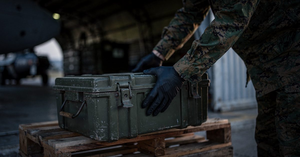 Close on gloved hands in the blue-hour light of a hangar doorway in South Korea, lowering an unused ammunition crate onto a pallet while a cropped shoulder in.