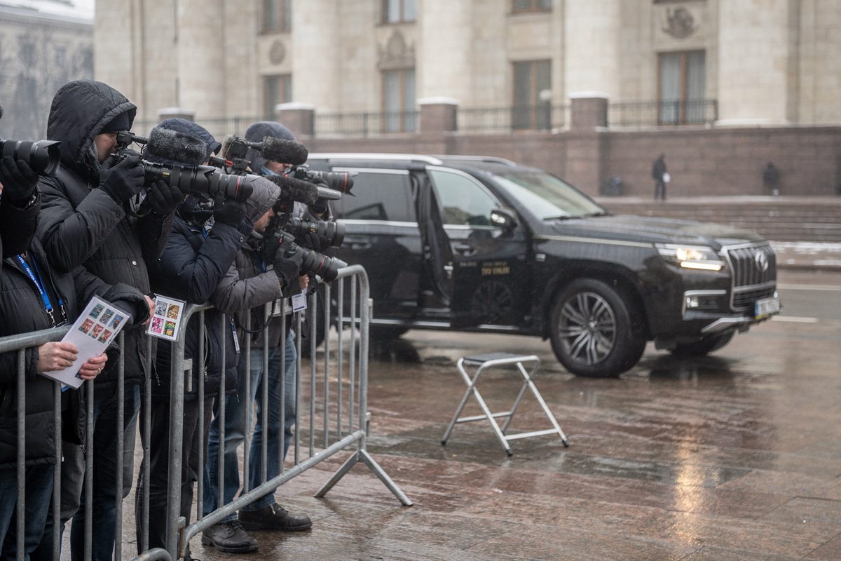Wide shot in flat pale afternoon light of a Kyiv government forecourt, journalists in dark parkas pressed behind a metal barrier as two black SUVs idle with a.