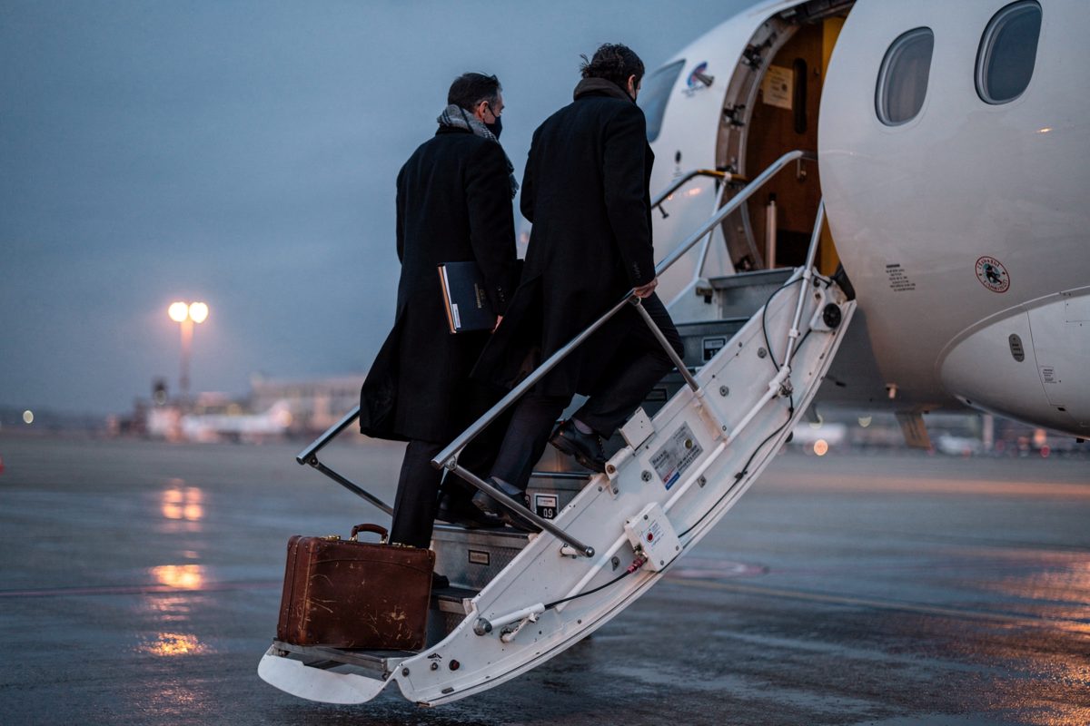 Medium shot at dusk on an airport apron, three aides in long overcoats climbing the stairs of an unmarked white jet, a leather document case propped upright.