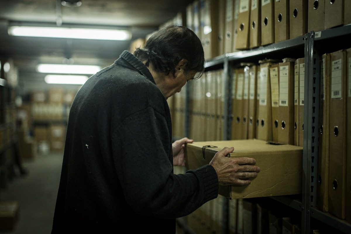 A dim basement records corridor lit by fluorescent strips with one tube dead, shot from behind a stooped archivist pulling an unmarked cardboard box file from.