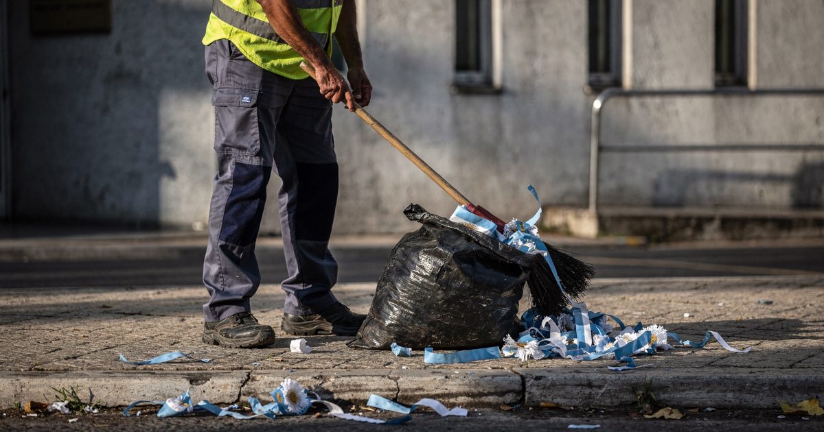 Harsh morning sunlight on a concrete municipal building as a worker seen from the waist down sweeps discarded blue and white paper bunting into a heavy black.
