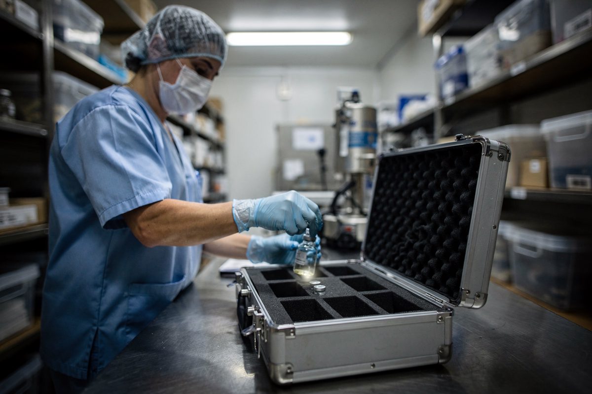 Fluorescent light in a clinical storage room as a technician in scrubs, face turned away, places a sealed glass vial containing a single drop of clear liquid.