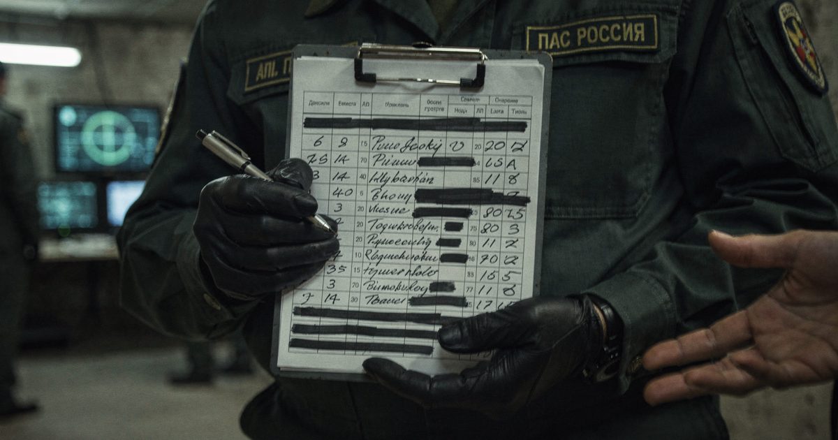 Close-up of a military officer's hands holding a clipboard with a heavily redacted, handwritten spreadsheet, standing under the harsh fluorescent lights of a.