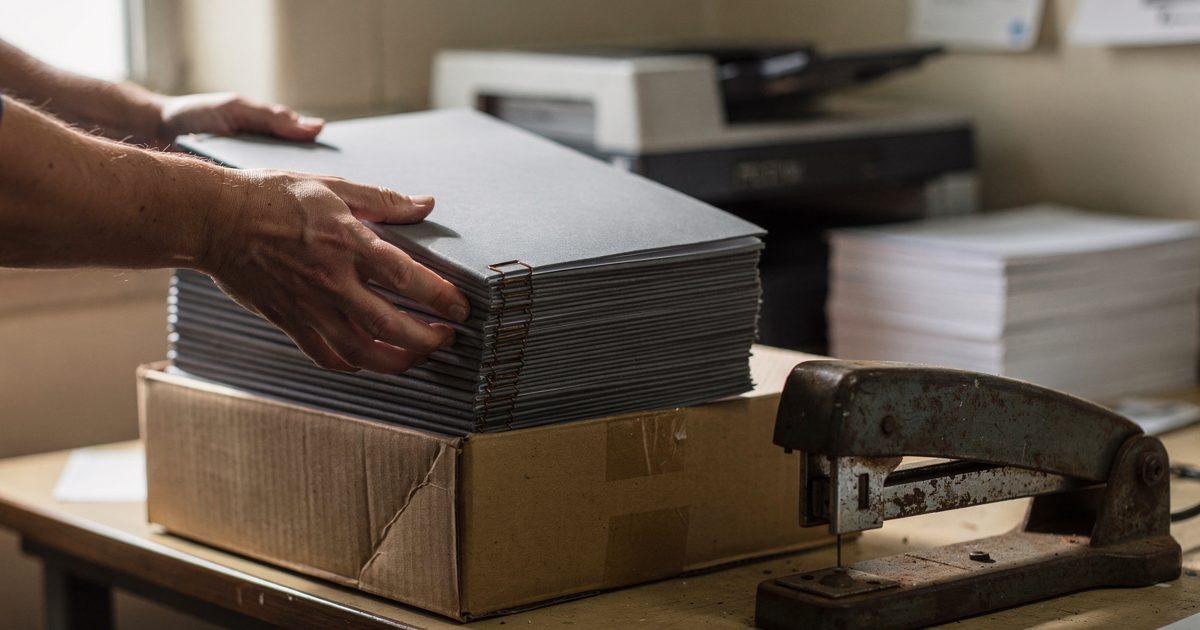 Close-up of two hands lifting a stack of identical grey stapled reports out of a cardboard box in an institutional print room, hard morning light from high.