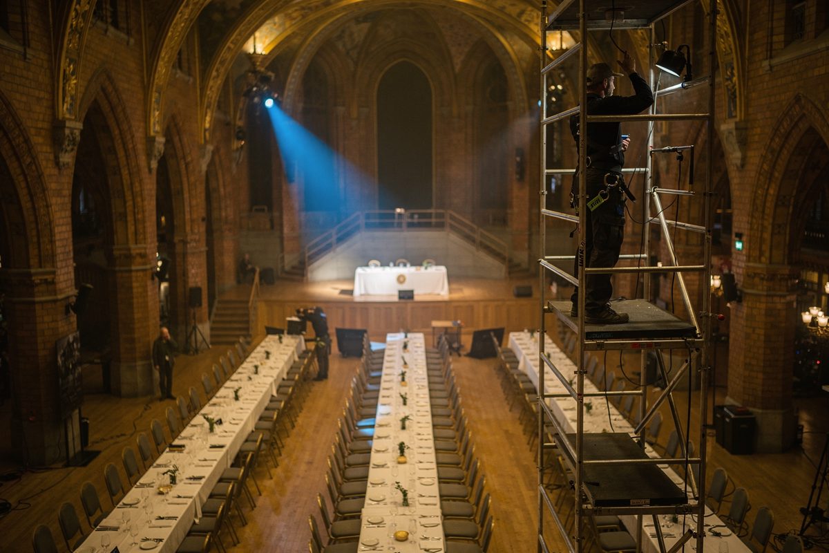 Wide shot across the gilded Blue Hall of Stockholm City Hall during a morning lighting rehearsal, long banquet tables laid for hundreds, a technician high on a.
