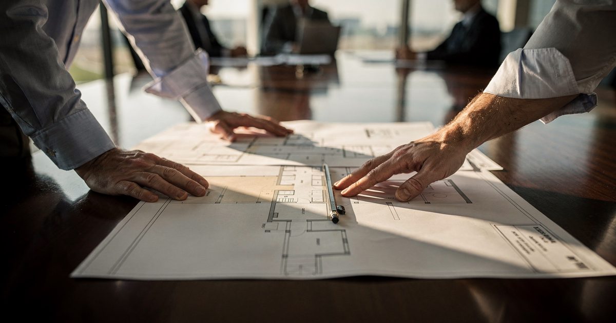 Close-up of two hands smoothing a large printed architectural floor plan across a polished conference table under warm late-afternoon light, a pencil resting.