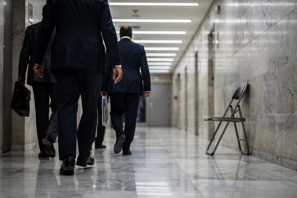 Wide shot from behind a line of part-cropped suited officials walking a marble Gulf ministry hallway at night, fluorescent strips overhead, a single folded.