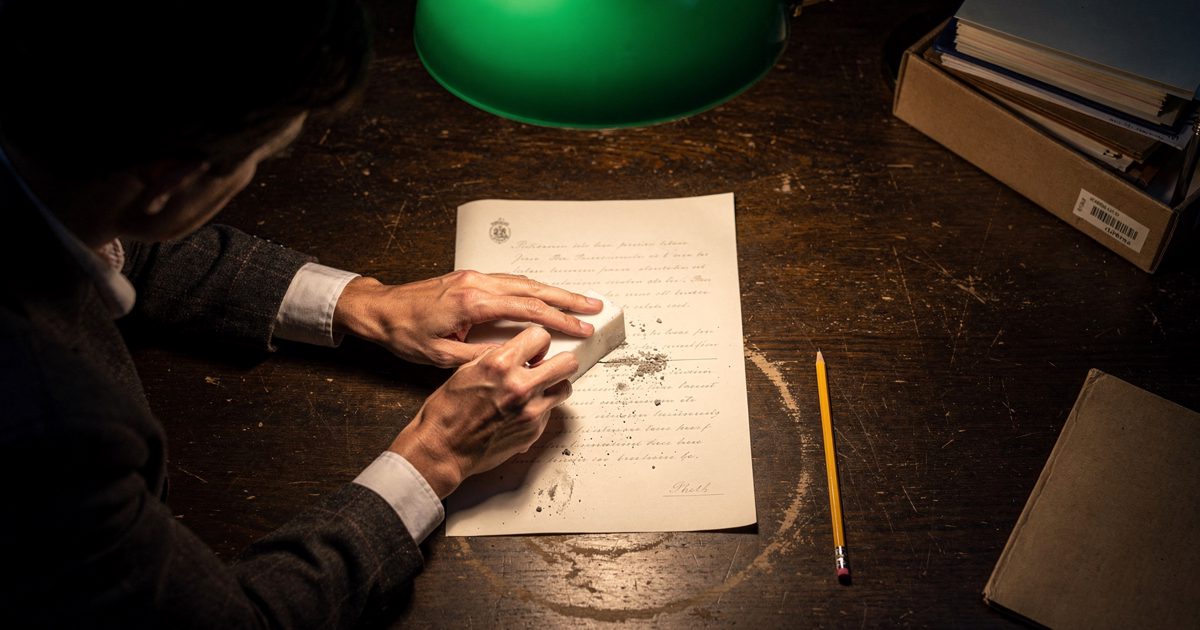 Overhead shot straight down onto a dark wooden archive table under a low green-shaded lamp late in the evening, two archivists leaning in from opposite edges.
