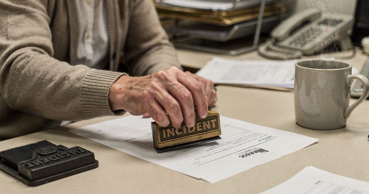 Close detail at desk level under flat fluorescent light in a windowless Ottawa government office, a hand in a cardigan sleeve pressing a wooden-handled rubber.