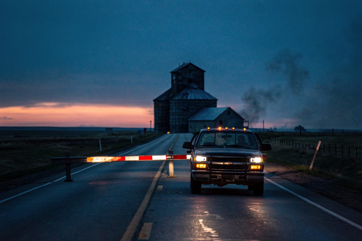 Wide shot at blue dusk of a two-lane prairie highway outside Wainwright, Alberta, a grain elevator silhouetted against the last orange band of sky and a lone.