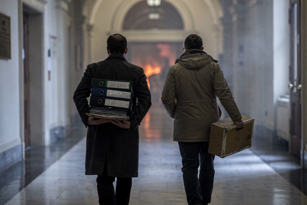 Medium shot from behind in a Parliament Hill corridor in cold early-morning light, two staffers in winter coats walking away down a long marble hallway, one.