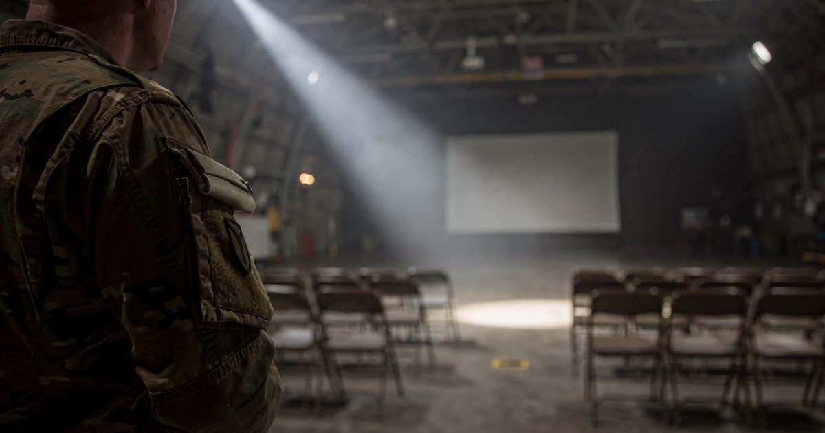 Close detail of a projector beam cutting through dusty air inside a vast military aircraft hangar at dusk, a soldier's cropped shoulder and folded cap in the.