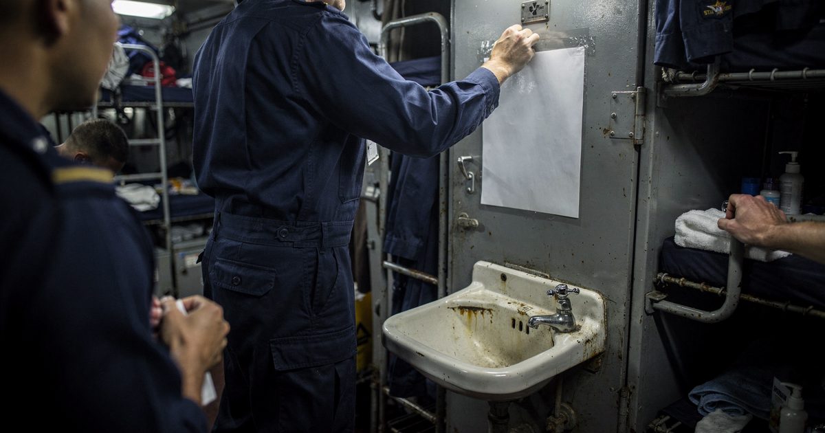 In a crowded aircraft-carrier berthing compartment at night under cold fluorescent tubes, a cropped figure in a navy working uniform tapes a single laminated.