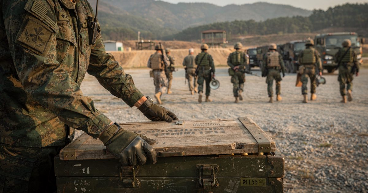 In warm late-afternoon light on a gravel Korean training range, gloved hands lower a lid onto a wooden crate stencilled for blank ammunition while a blurred.