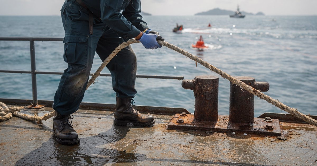 Harsh midday sun glaring off the wet metal deck of a docked maritime patrol boat, a crew member shot from the waist down securing a thick nylon mooring line to.