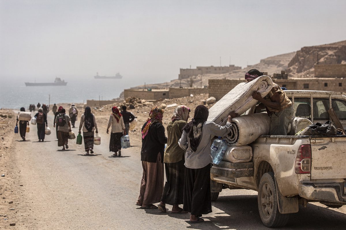 Wide shot in hard midday glare on a dusty coastal road in western Yemen, a dozen people seen from behind loading mattresses, blankets and plastic water cans.