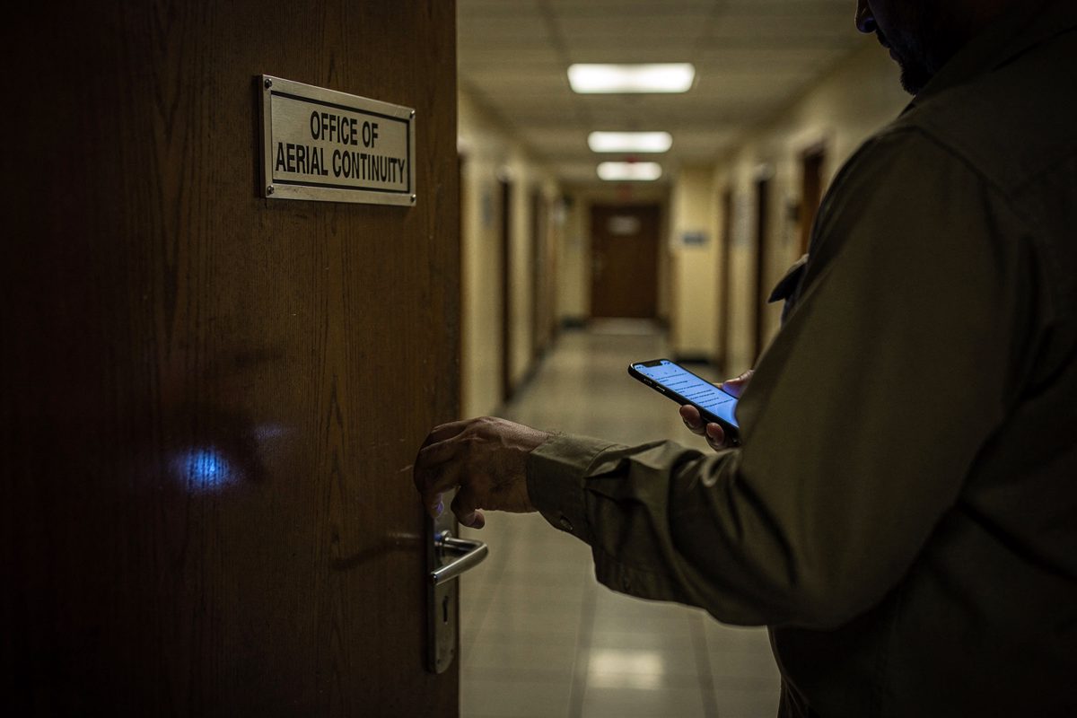 In a dim government corridor at dusk, a person in a khaki shirt cropped at the shoulder pushes open a door with a plaque reading "Office of Aerial Continuity".