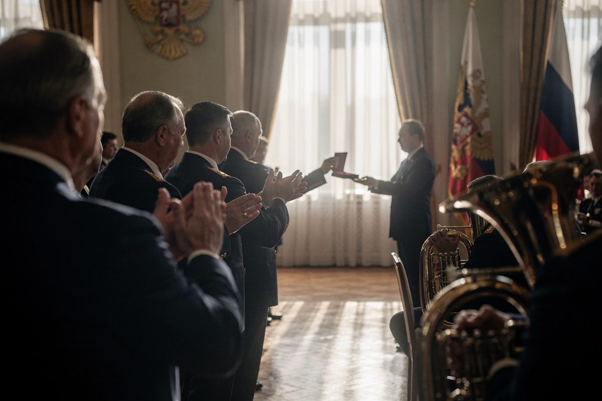 Wide shot in low, hard winter sun slanting through tall Kremlin hall windows late in the afternoon, a line of officials in dark suits seen from behind.