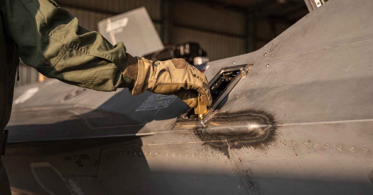 Close detail in hard late-afternoon hangar light of a gloved hand slotting a small brass key into a panel beside a fighter jet's rear canopy rail, the metal.