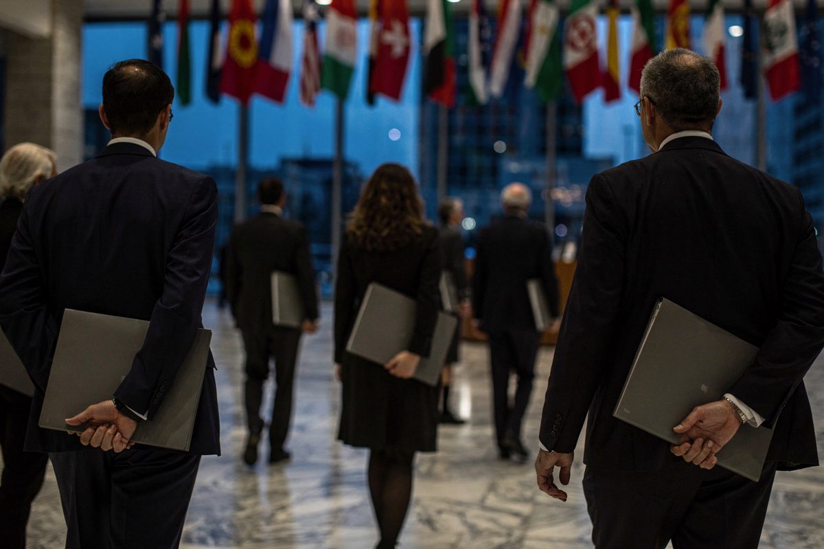 Wide shot from behind of delegates crossing the marble lobby of an international headquarters at dusk, member-state flags hanging overhead, each figure.