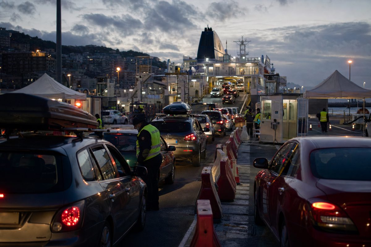 A dusk news photograph of cars waiting at a Mediterranean ferry terminal checkpoint, temporary barriers and inspection booths in view, officials in reflective.