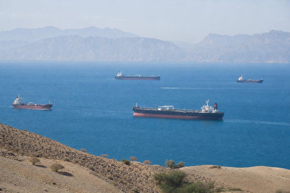 Documentary-style photograph of cargo vessels and oil tankers passing through a broad sunlit maritime strait, viewed from a distant shoreline with hazy.