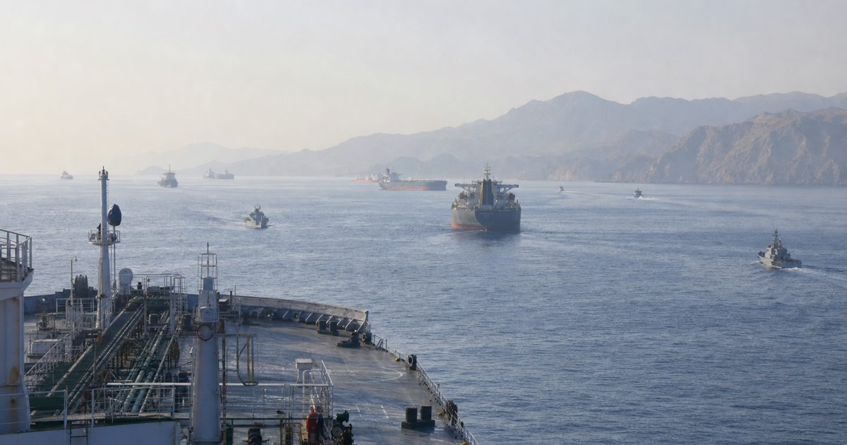 Wide editorial news photograph from the deck of a commercial vessel in the Strait of Hormuz, showing distant tankers, patrol craft silhouettes and a hazy.