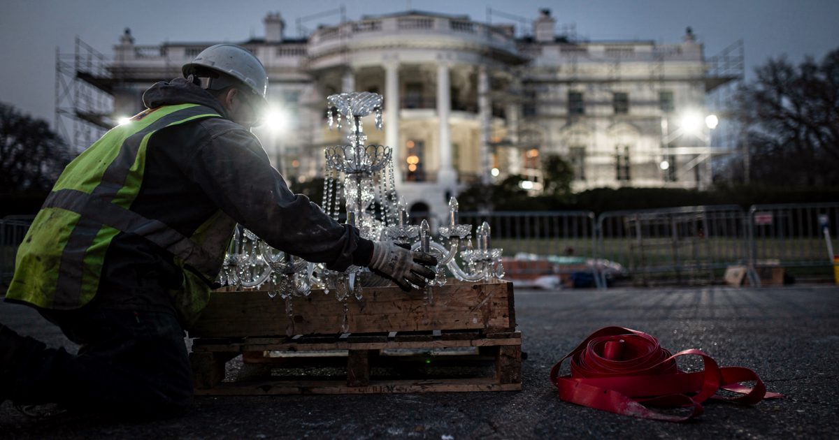 Dusk floodlights rake White House East Wing scaffolding as two workers, seen from behind and cropped at the shoulders, ease a crate of crystal chandelier arms.