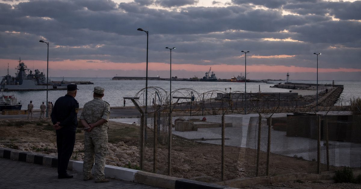 A wide documentary news photograph of a Mediterranean naval facility at dusk, with concrete piers, distant ships, security fencing and anonymous uniformed.