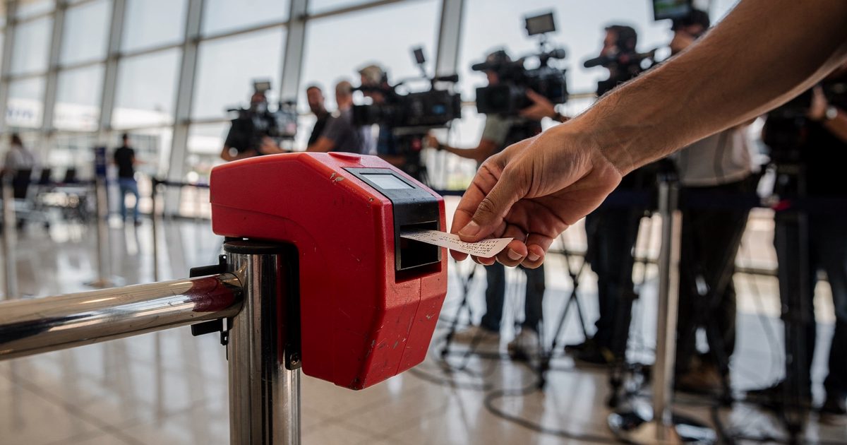 Close detail at waist height in bright midday sun pouring through the glass wall of an airport arrivals hall, a hand pulling a paper number from a red.