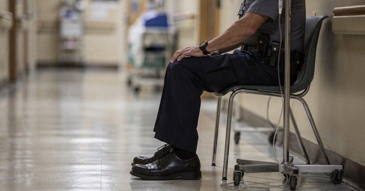 Medium shot from the corridor of a quiet hospital ward under harsh fluorescent light at midday, a uniformed state trooper seen from the waist down sitting on a.