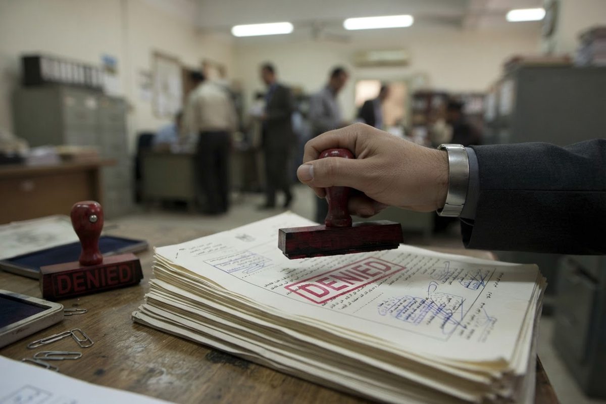 A close-up of a generic mid-level bureaucrat's hand decisively stamping a thick stack of meaningless paperwork with a heavy, red 'DENIED' stamp.