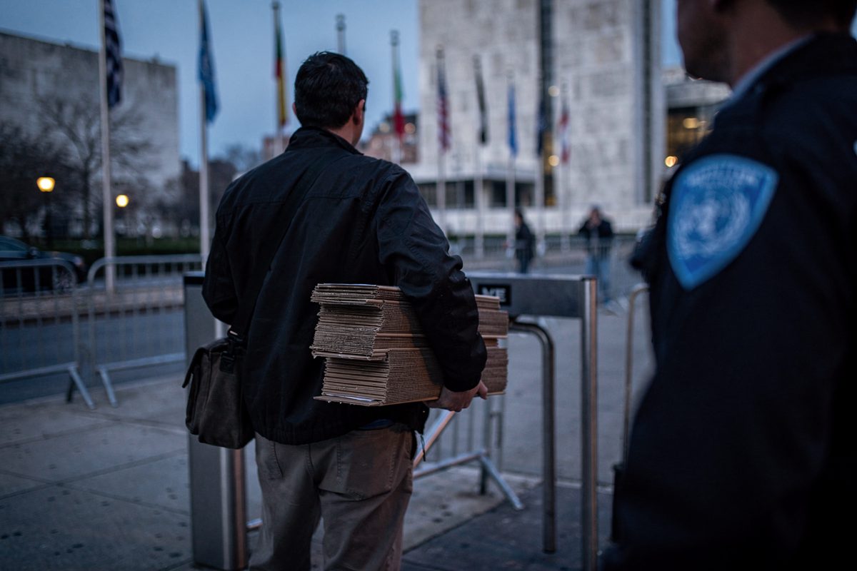 Mid-shot at blue dawn on the First Avenue sidewalk outside UN headquarters, a courier seen from the back waiting at the visitor turnstile with a stack of.