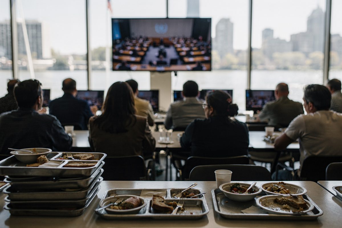 Wide shot at hard noon inside the UN headquarters cafeteria, diners seen from behind at long tables watching a muted screen that shows a blurred assembly hall.