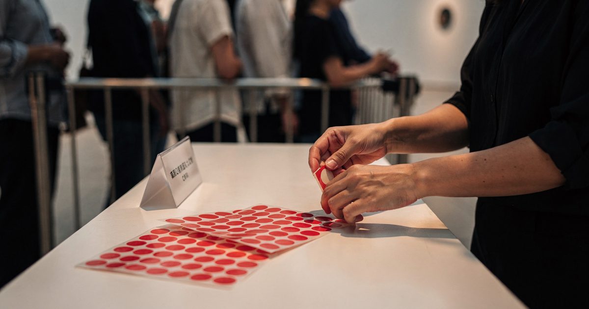 Close early-evening light on a museum admissions desk as an attendant's hands peel red adhesive circles from a backing sheet beside a printed card reading.