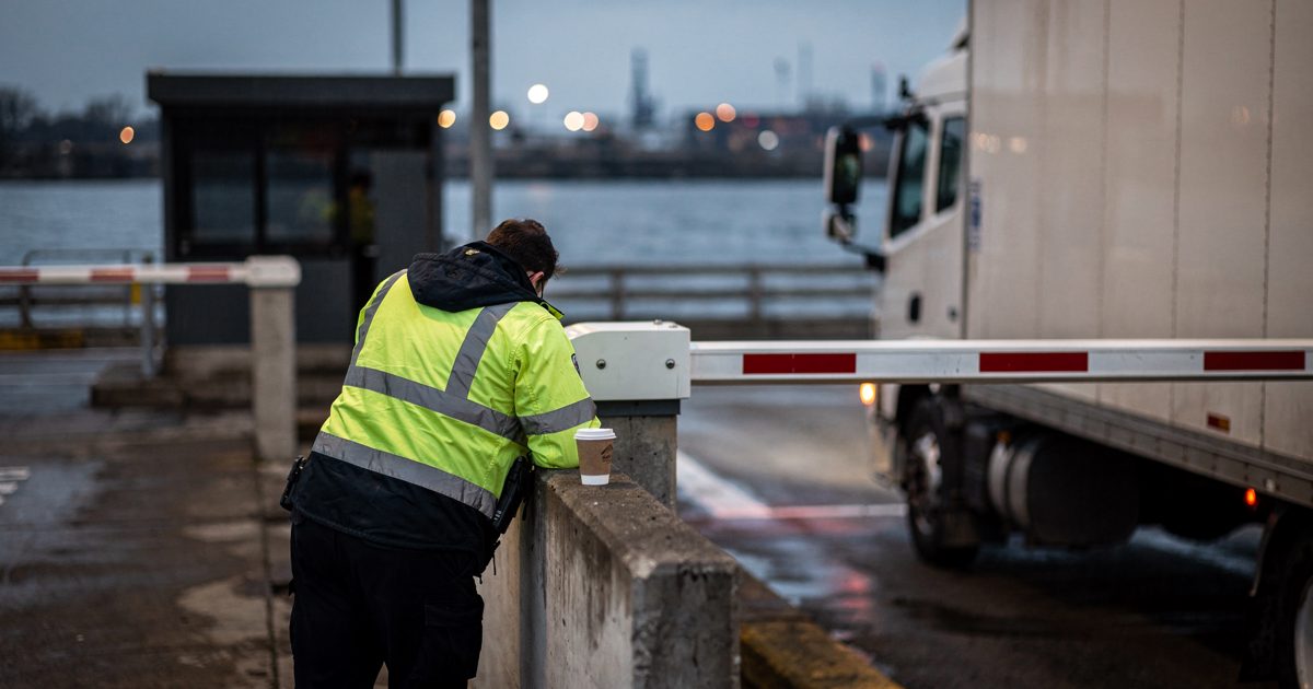 A solitary customs officer in a high-visibility jacket leans against a closed toll boom at dusk, a half-empty cup of coffee resting on the concrete barrier.