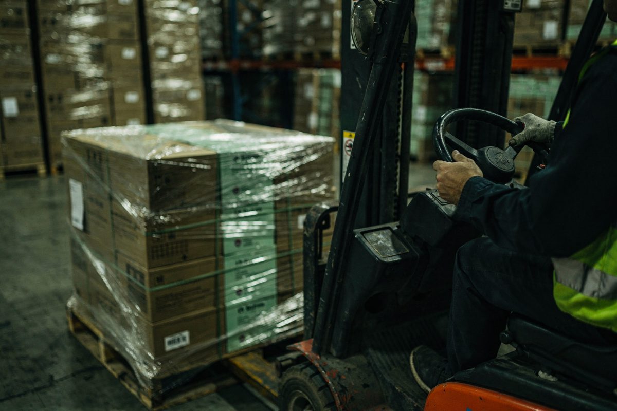 A forklift operator's gloved hands grip the steering wheel inside a warehouse under harsh fluorescent lights, a pallet of shrink-wrapped cardboard boxes.