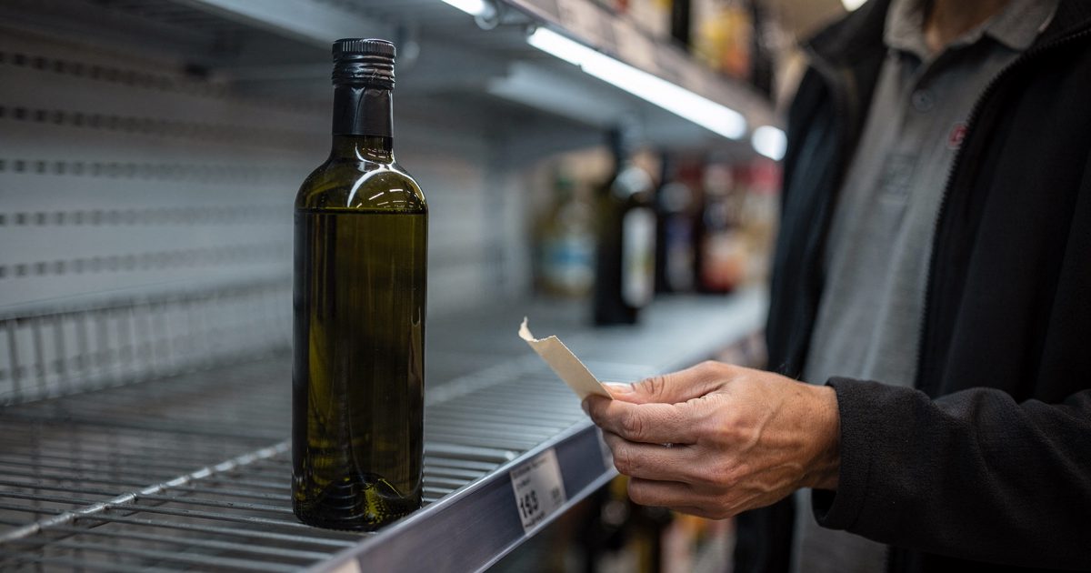 Close detail under harsh supermarket fluorescent light of an unlabelled dark glass oil bottle on a half-empty grocery shelf, a shop worker cropped at the.