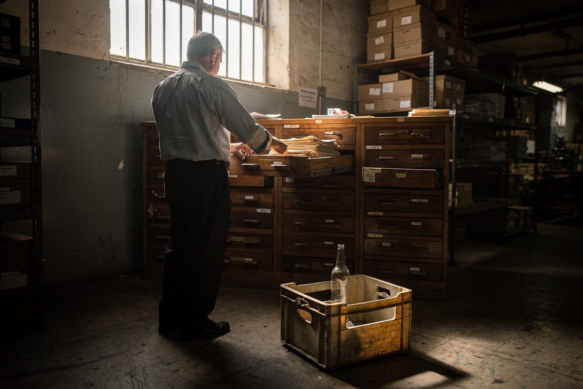 Late afternoon light from a high barred window in a basement records room of a former tax office, a civil servant seen from behind flipping through a wooden.