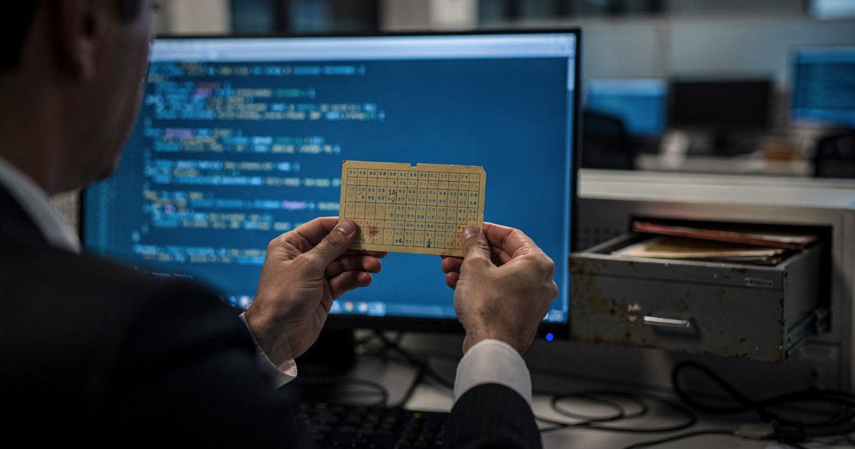 Close-up under the cold blue glow of a monitor in a darkened open-plan software office at night, a pair of hands in shirt cuffs holding a yellowed cardboard.