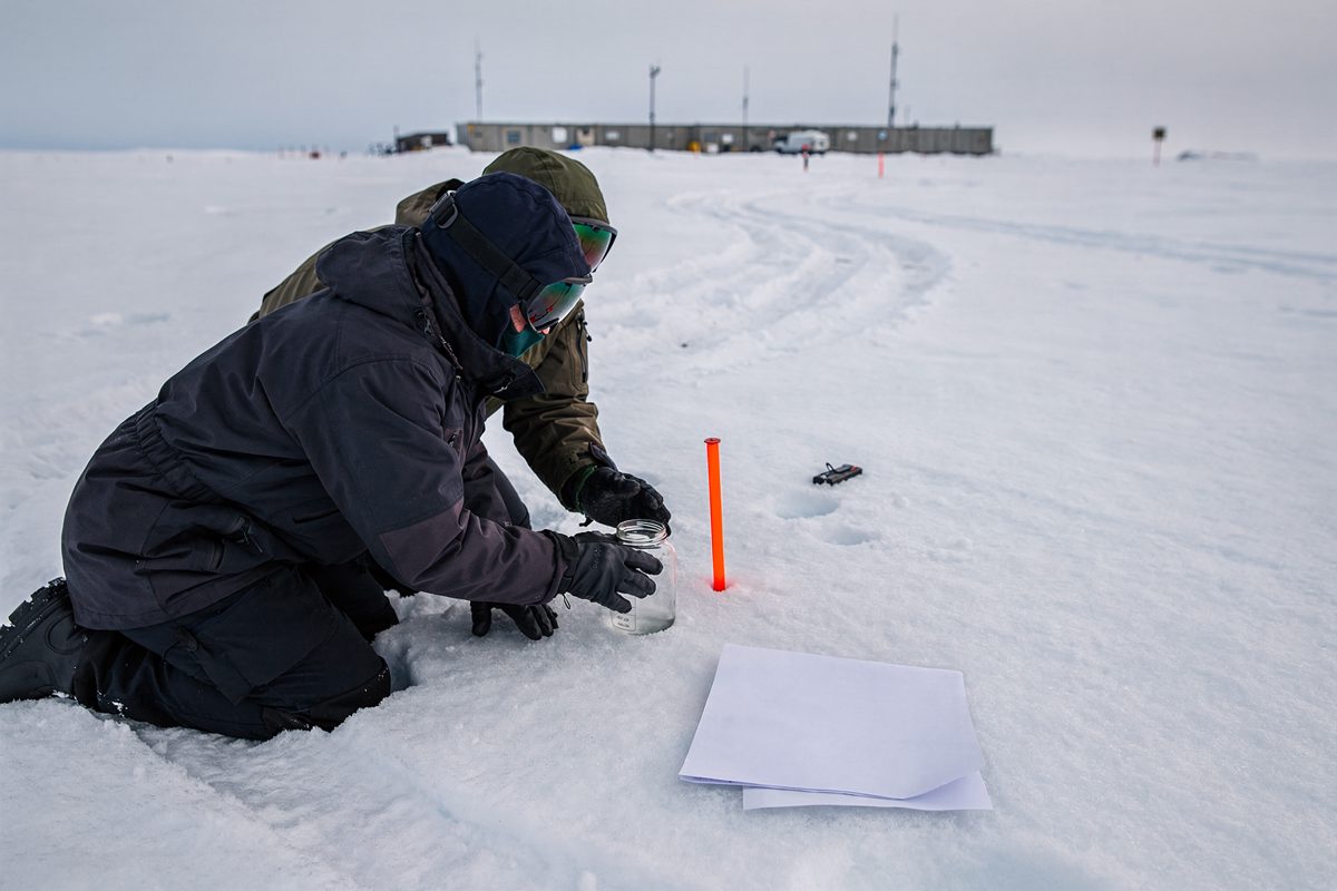 Wide shot in flat polar daylight on the inland ice, a survey team cropped at the shoulders kneeling to press a clear jar of water into the snow next to an.