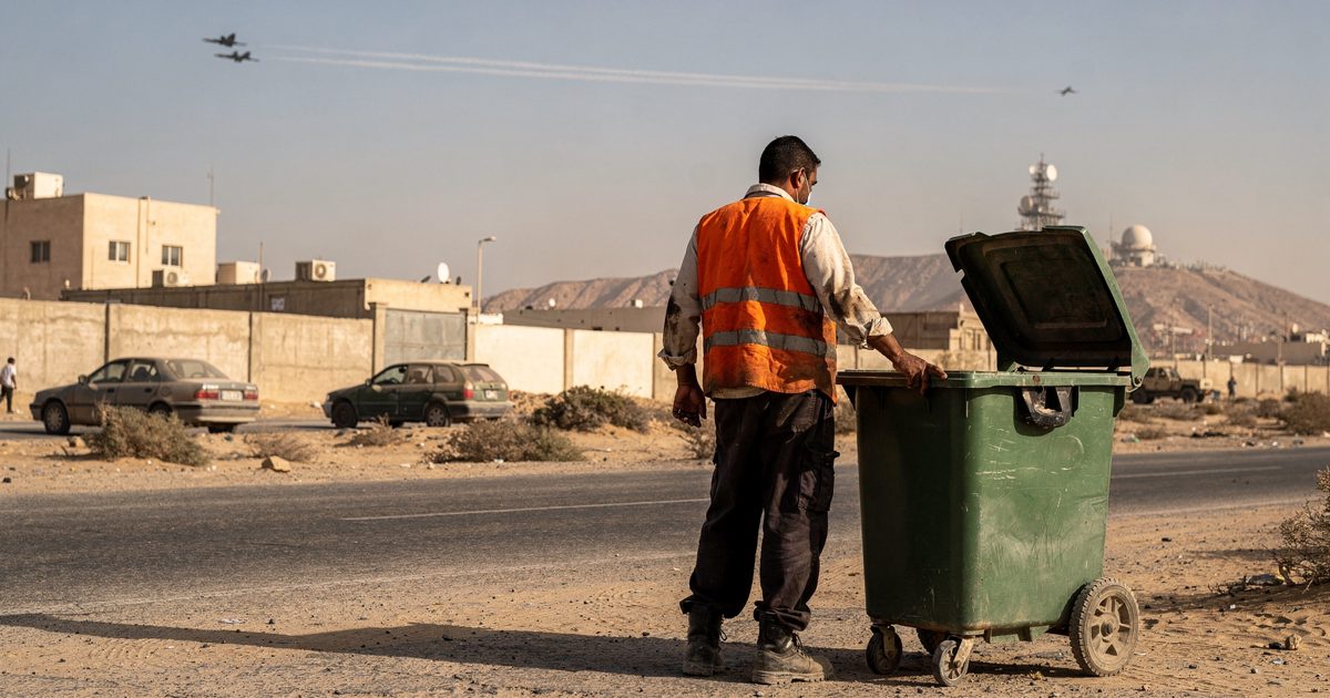 A municipal worker in a faded orange high-visibility vest stands idle beside an empty green waste bin on a dusty roadside in Muscat under harsh morning.